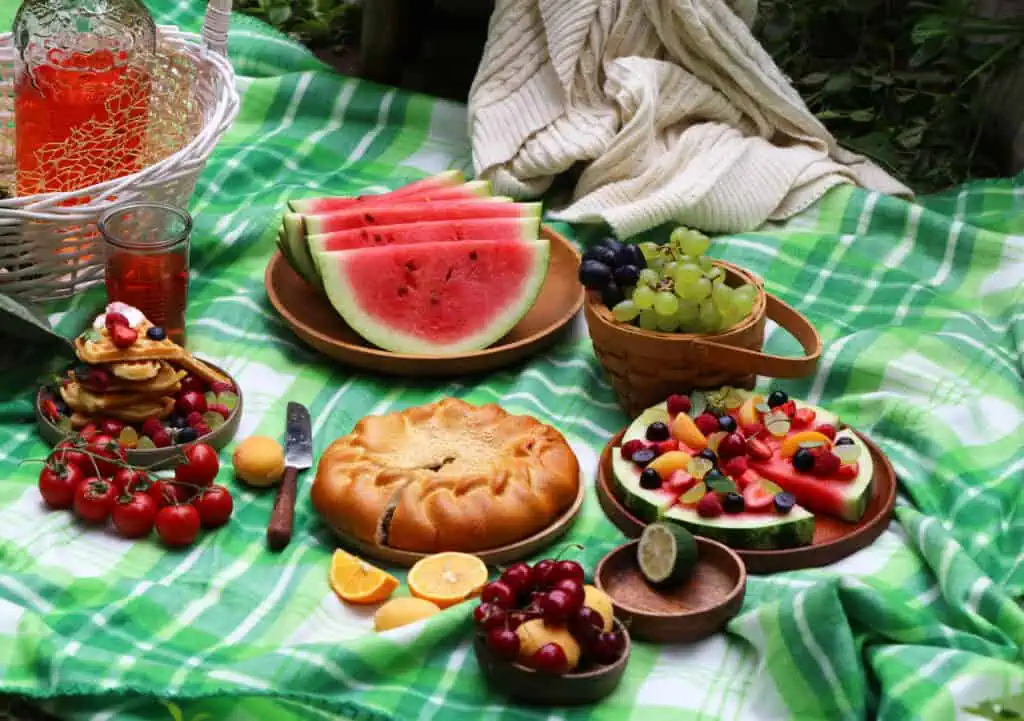 A picnic spread on a green checkered blanket with watermelon slices, fruit, pie, crackers, grapes, cherries, tomatoes, citrus slices, and a pitcher of red drink.