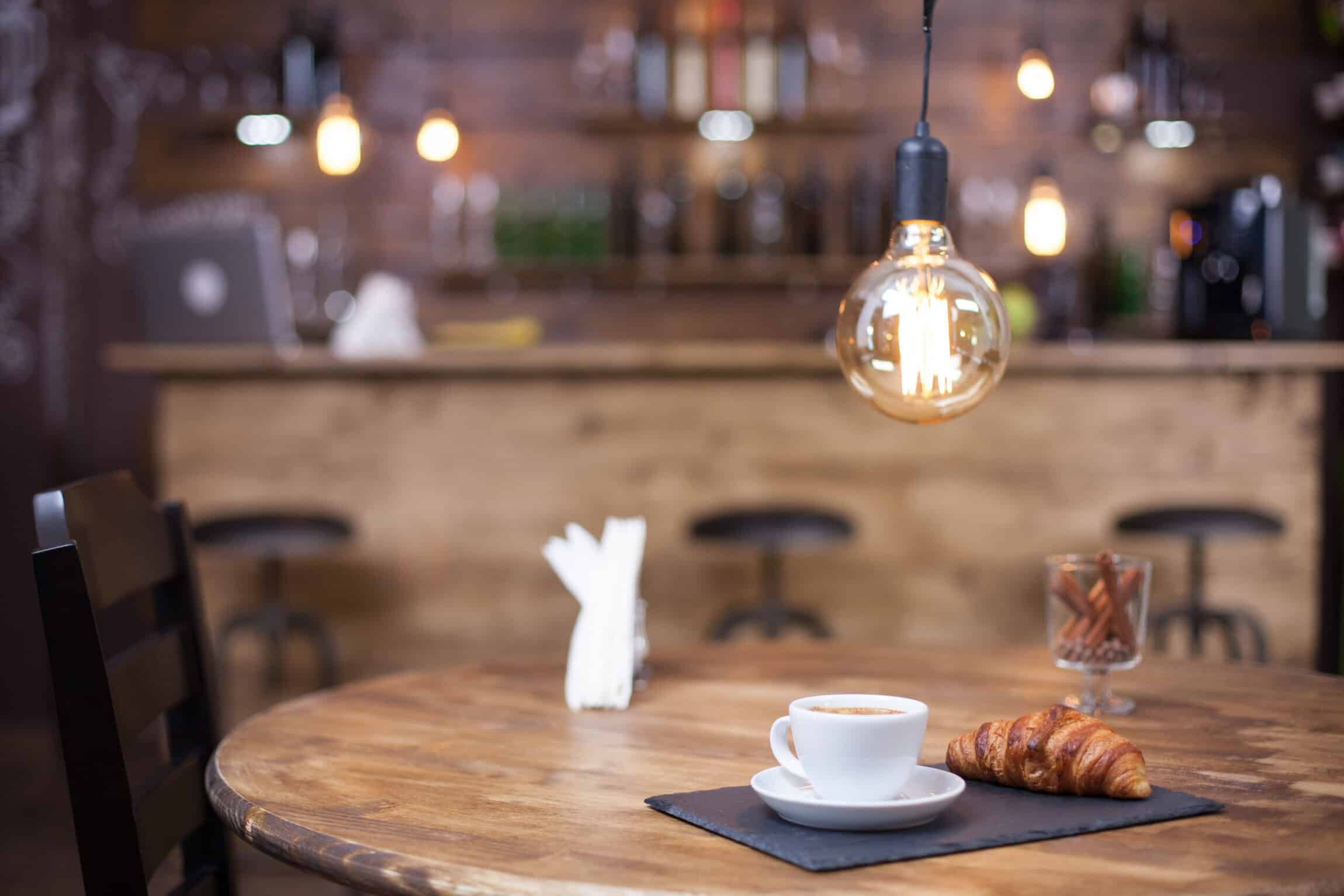 A cup of coffee and a croissant on a wooden table in a cozy café with a blurred background featuring bar stools and warm lighting.