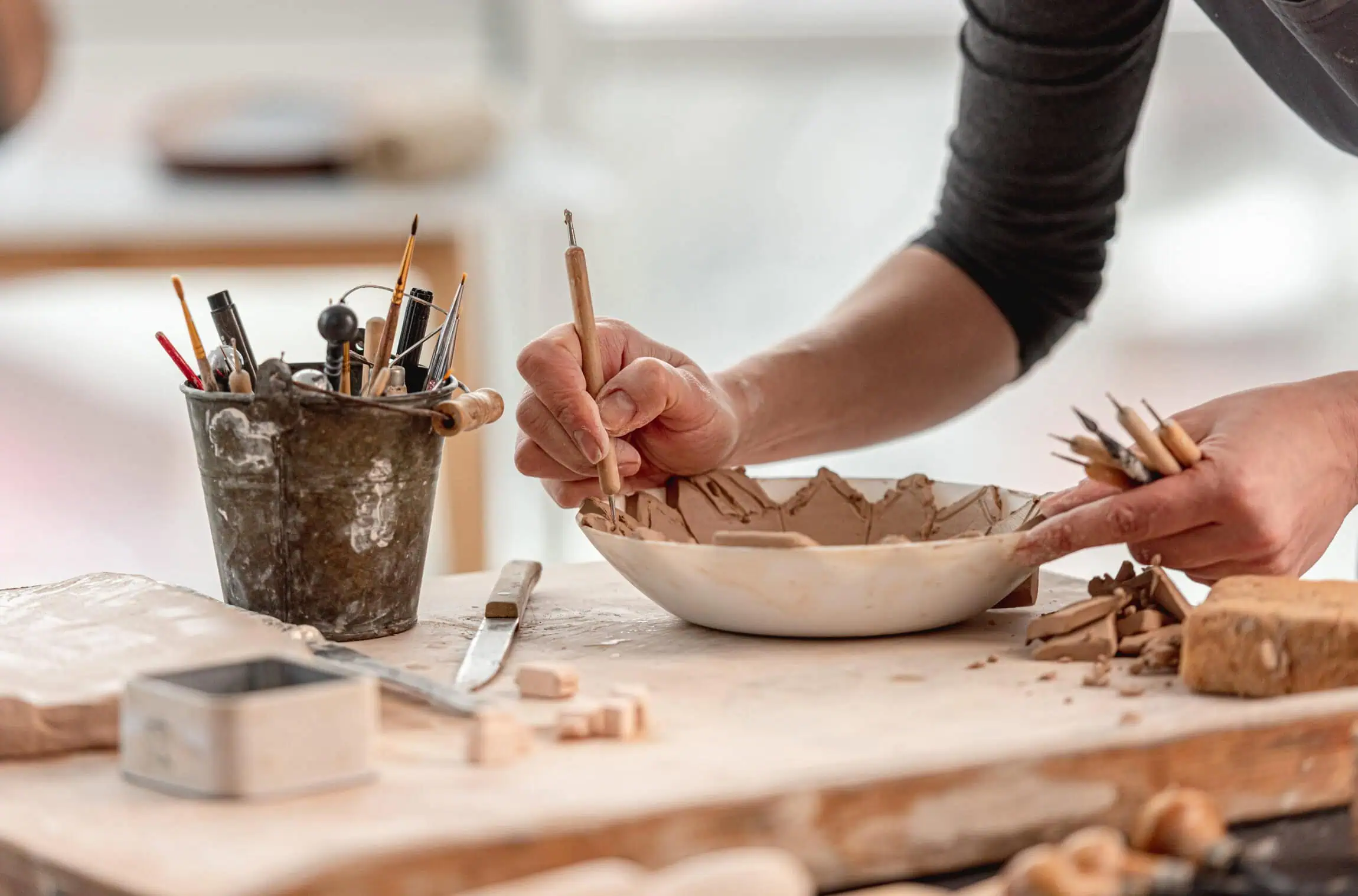 Person carving patterns into a clay bowl with sculpting tools; various pottery tools and clay pieces are on the worktable.