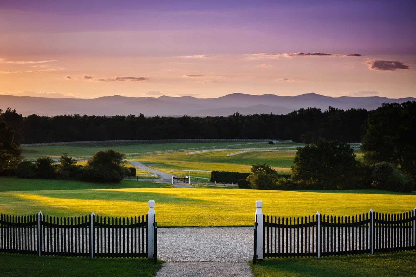 View of a fenced walkway leading to open grassy fields and winding paths, with trees and mountains in the background under a colorful sunset sky.