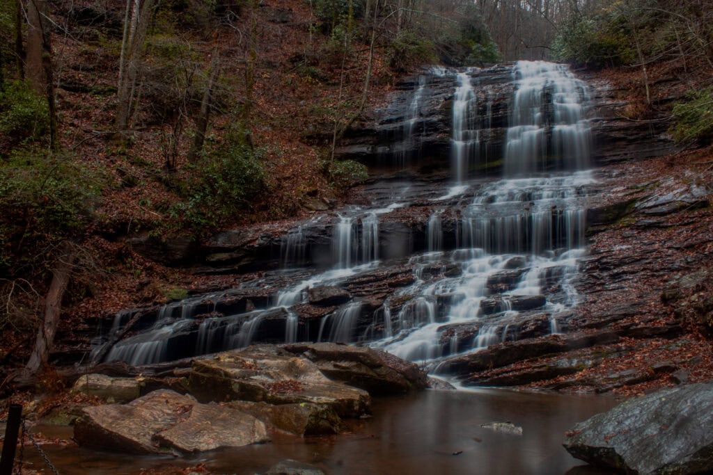 A tiered waterfall cascades over dark rocks in a forested area with bare trees and scattered greenery, surrounded by wet leaves and boulders.