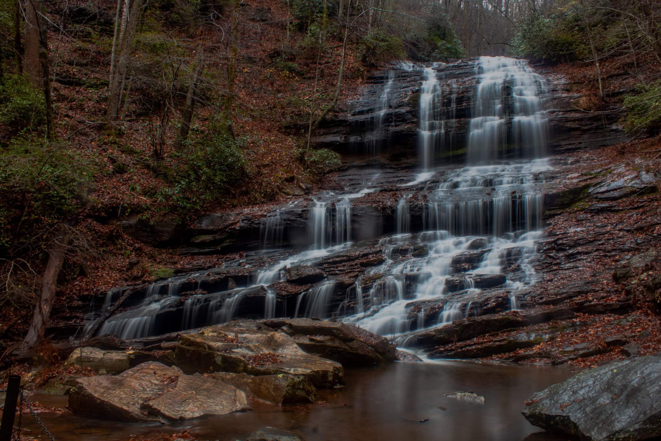 A tiered waterfall cascades over dark rocks in a forested area with bare trees and scattered greenery, surrounded by wet leaves and boulders.