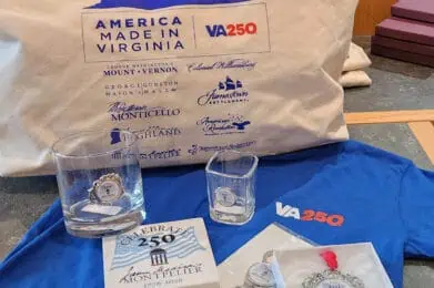 A VA250 tote bag, T-shirt, glassware, coaster, keychain, and ornament, all featuring "America Made in Virginia" and Monticello 250th anniversary branding, displayed on a table.