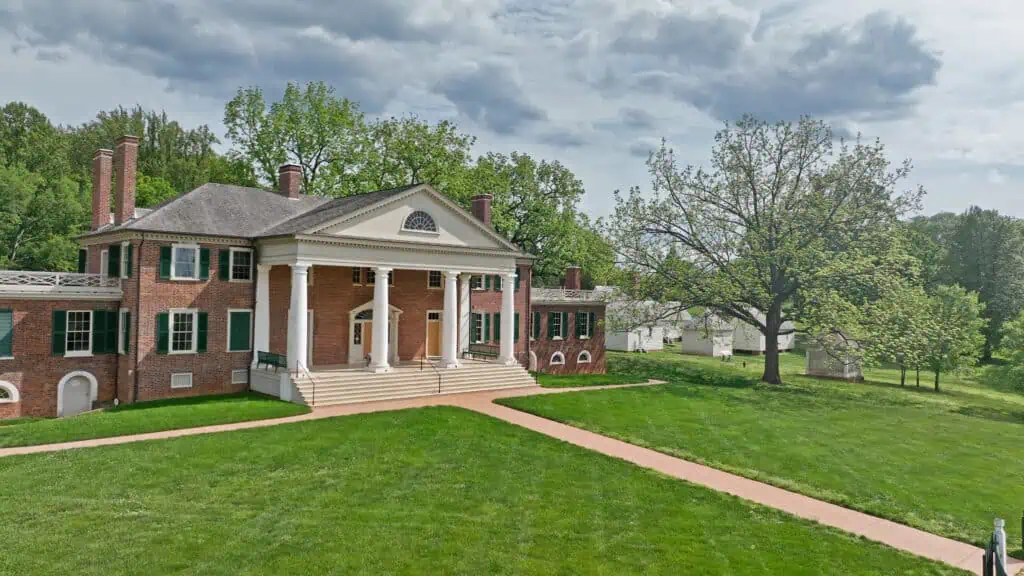 A large red-brick mansion with white columns, green shutters, and a broad lawn, surrounded by trees under a cloudy sky.