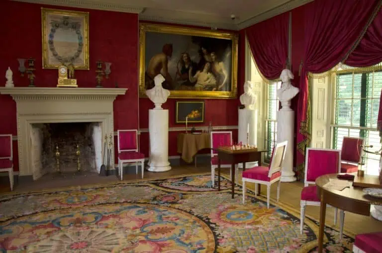 A formal room with red walls and drapes, ornate rug, fireplace, classical busts on pedestals, paintings, and elegant furniture arranged around a chess table.