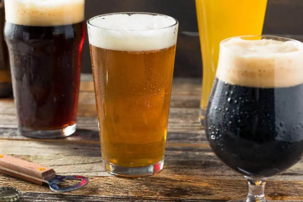 Four glasses of different types of beer, including pale, amber, and dark varieties, are placed on a wooden table with a bottle opener and bottle cap nearby.