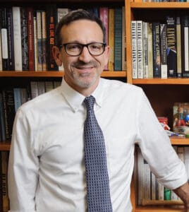 A man wearing glasses, a white shirt, and a patterned tie stands in front of a bookshelf filled with books.