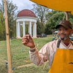 A man in an apron stands under a yellow tent, holding a small object, with a white domed pavilion and trees visible in the background.