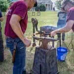 Two people wearing maroon shirts work at an outdoor blacksmith station, using an anvil and tools near a grill, with trees and a building in the background.