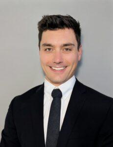 A man in a black suit, white shirt, and black tie is smiling at the camera against a plain light gray background.