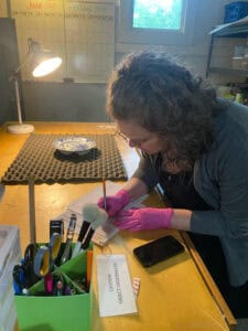 A woman wearing pink gloves uses a brush and tool to clean or examine an object on a desk, with supplies, a phone, and a "Caution: Unlabeled Objects" sign nearby.