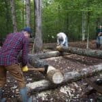Several people outdoors in a wooded area use axes to shape and construct a log structure on the forest floor.