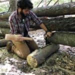 A man in outdoor clothing kneels on the ground, using a wooden mallet and chisel to carve a log, with stacked logs and forest in the background.