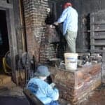 Two workers repair a brick fireplace and chimney inside a dim, rustic building. One stands on the hearth, applying mortar, while the other crouches nearby.