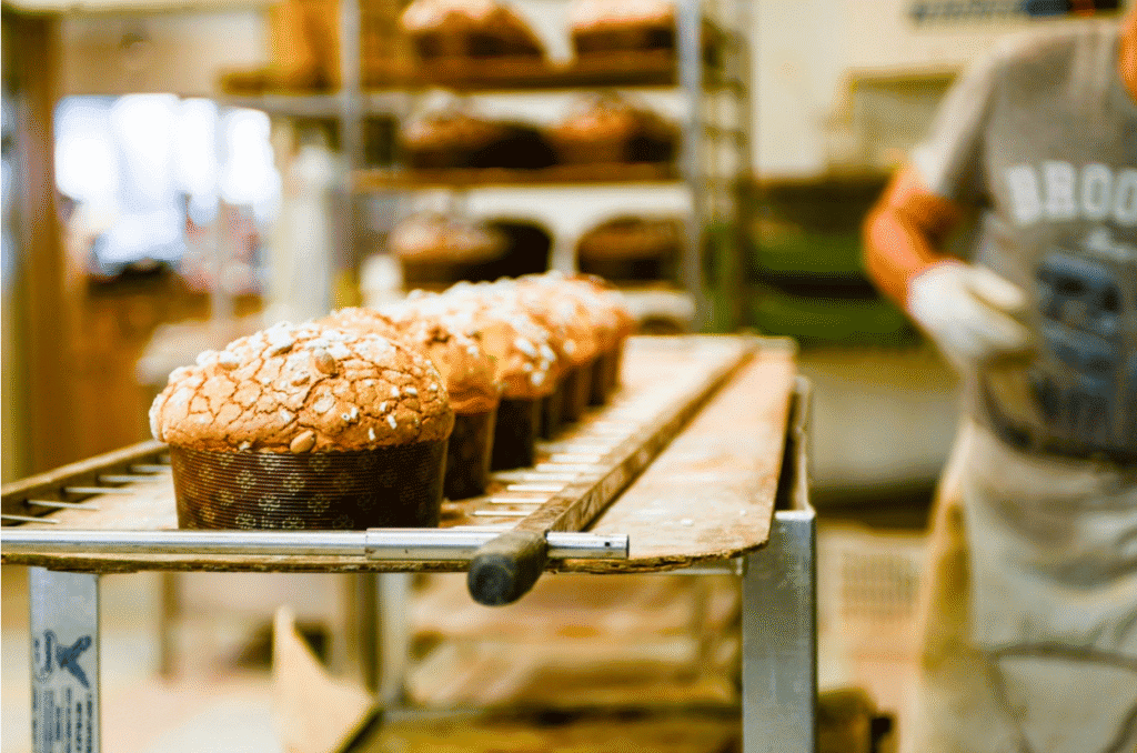 Rows of freshly baked panettone cool on a rack in a bakery, with a worker in an apron standing nearby.