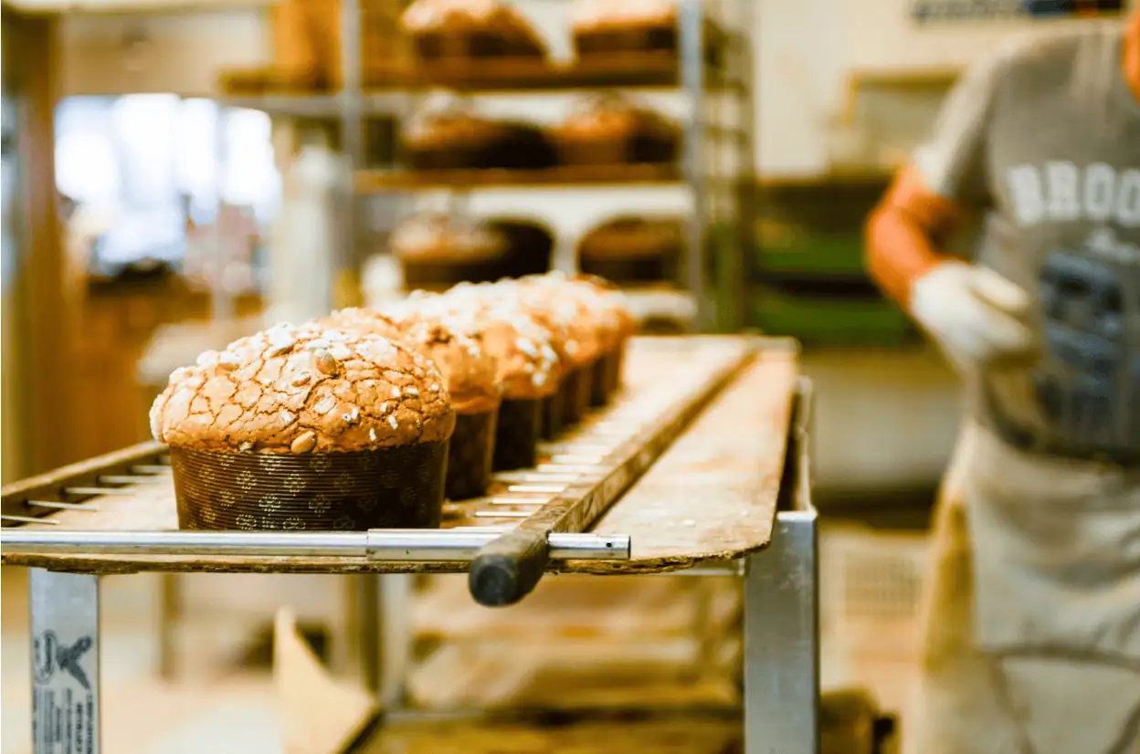 Rows of freshly baked panettone cool on a rack in a bakery, with a worker in an apron standing nearby.