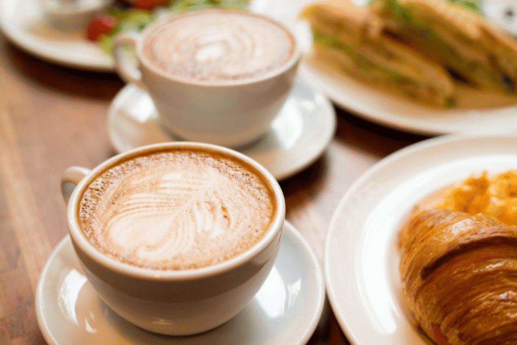 Two cups of cappuccino on saucers sit on a table beside plates of a croissant, scrambled eggs, and a sandwich.