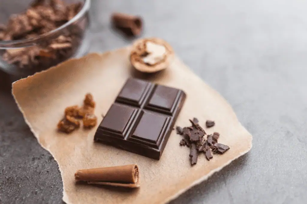 A square of dark chocolate on parchment paper, surrounded by chocolate shavings, a chocolate curl, some nuts, and small pieces of dried fruit.