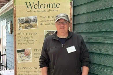 A man stands in front of a green building next to a "Welcome to the Archaeology Laboratory" sign with information about the lab’s activities.