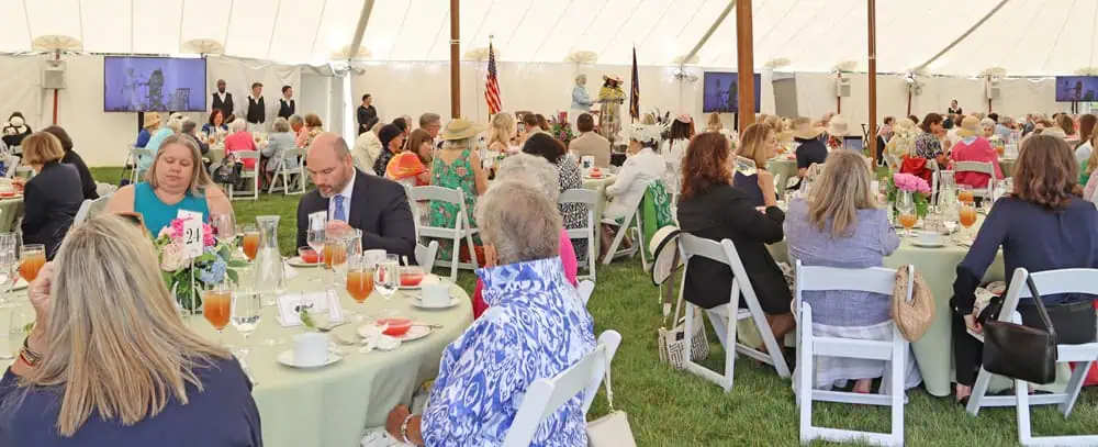 People seated at round tables under a large white tent during a formal event, with floral centerpieces and drinks on the tables.