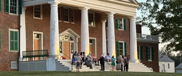 A group of people gathers and sits on the steps of a large brick building with white columns and green shutters in the evening.
