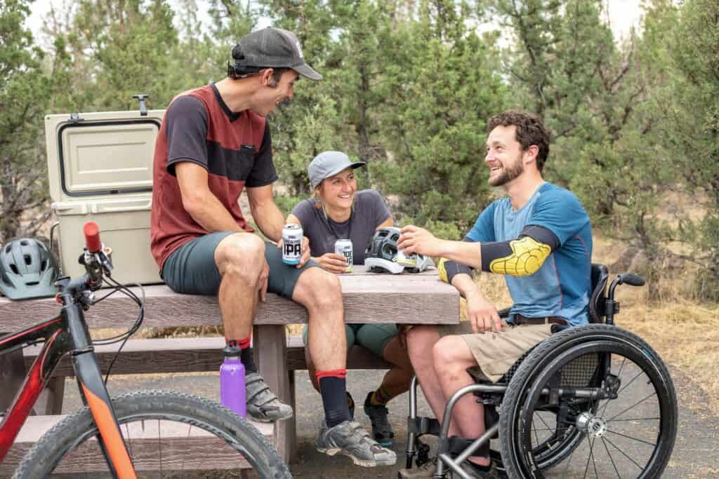 Three friends relax outdoors at a picnic table after biking, one using a wheelchair and all sharing drinks and helmets, surrounded by trees and adaptive gear.