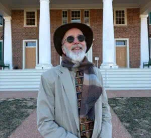 An older man with a white beard, wearing a hat, sunglasses, a tan jacket, a patterned tie, and a scarf, stands smiling in front of a historic brick building with white columns.