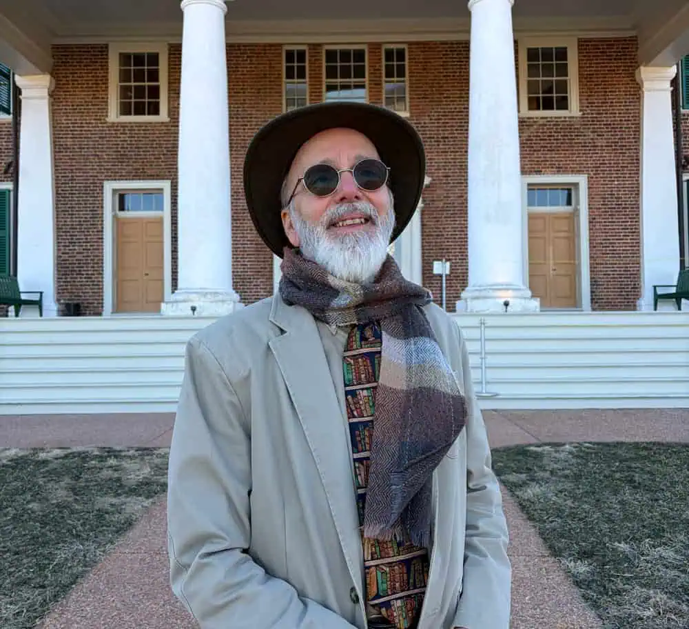 An older man with a white beard, wearing a hat, sunglasses, a tan jacket, a patterned tie, and a scarf, stands smiling in front of a historic brick building with white columns.