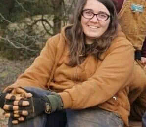 A person wearing glasses, a brown hoodie, and work gloves sits outdoors on a wooden surface, smiling at the camera. Another person in overalls is partially visible beside them.