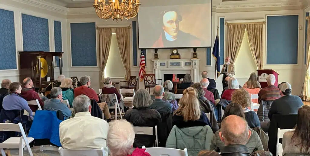 A group of people sits facing a speaker in a formal room as a portrait of a historical figure is projected on a screen at the front.