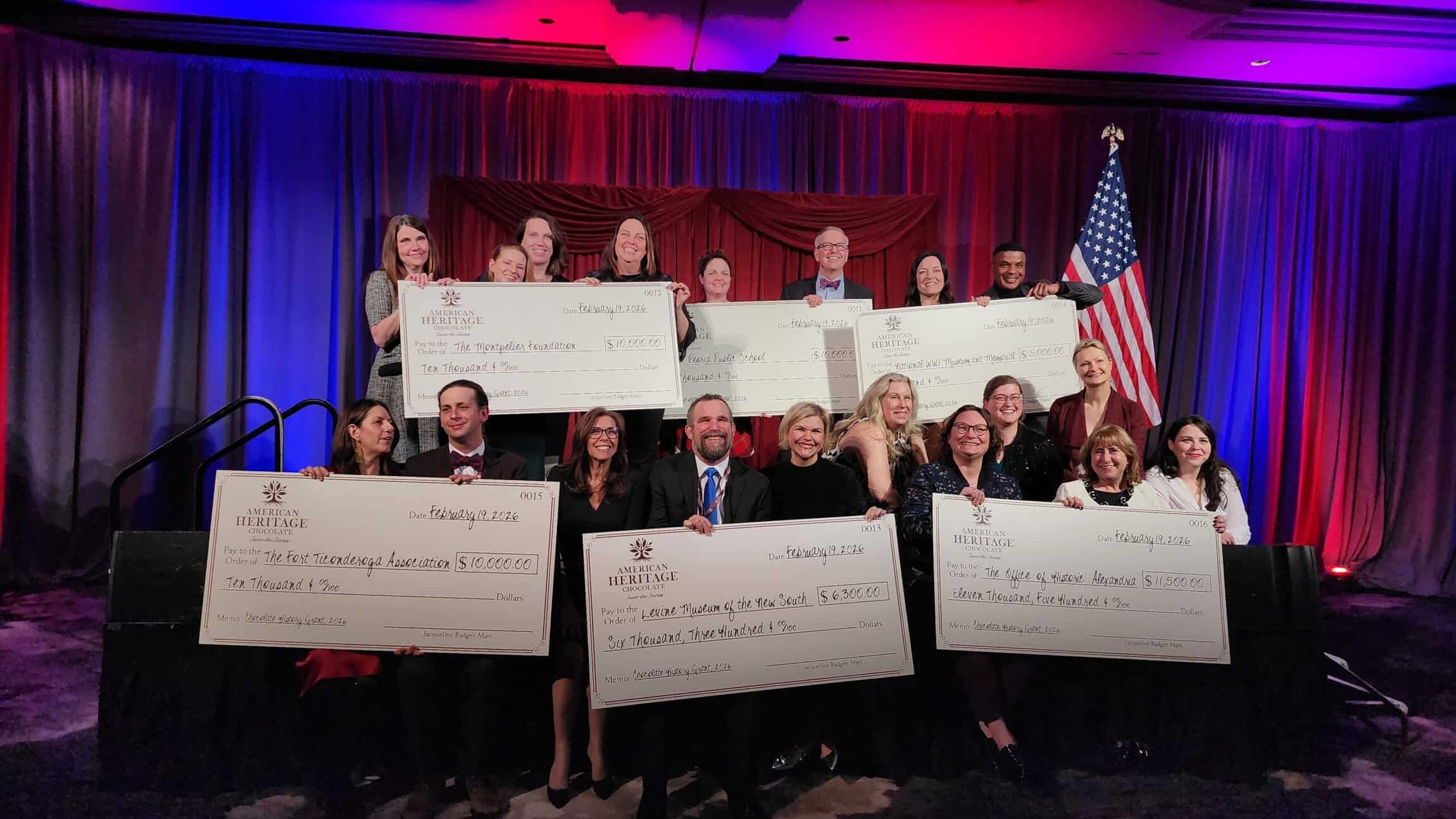 A group of people on stage holding oversized donation checks, with a U.S. flag and red, white, and blue lighting in the background.