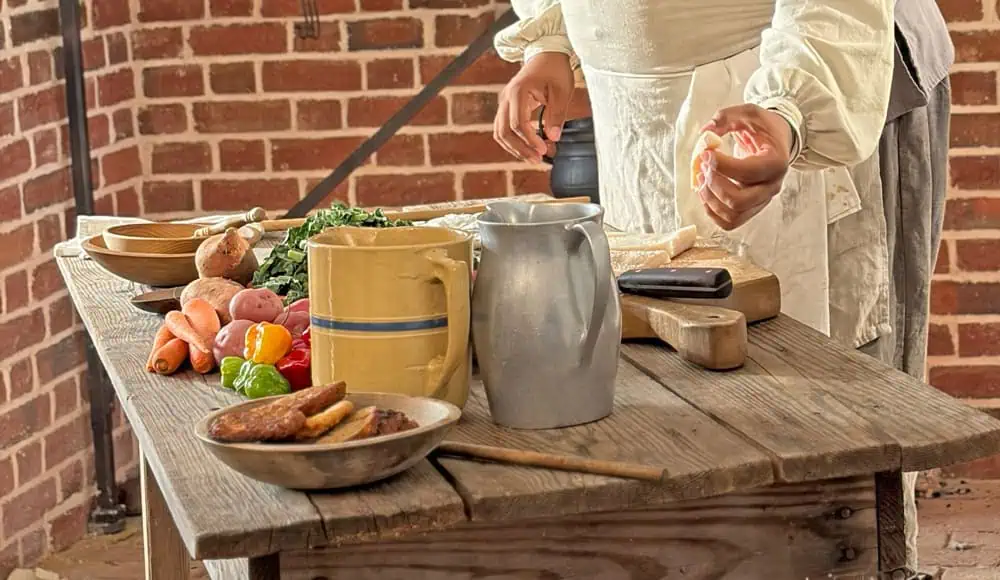 A person in historical clothing prepares food on a wooden table with vegetables, a ceramic bowl, a metal pitcher, and a plate of cooked sausages in a brick-walled kitchen.