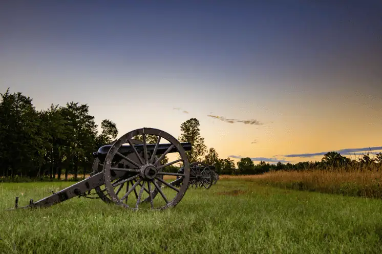 Civil War-era cannons lined up in a grassy field at sunset, with trees in the background and a soft orange and blue sky overhead.