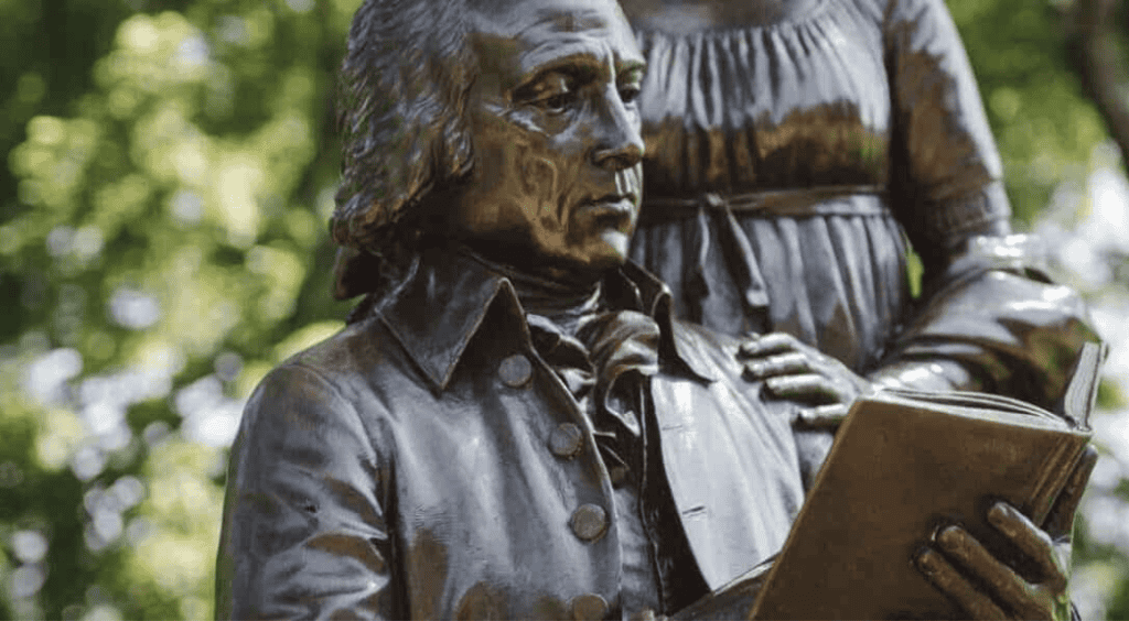 Bronze statue of James Madison in colonial-era attire reading a book, with a woman standing behind him, set in a green park setting.