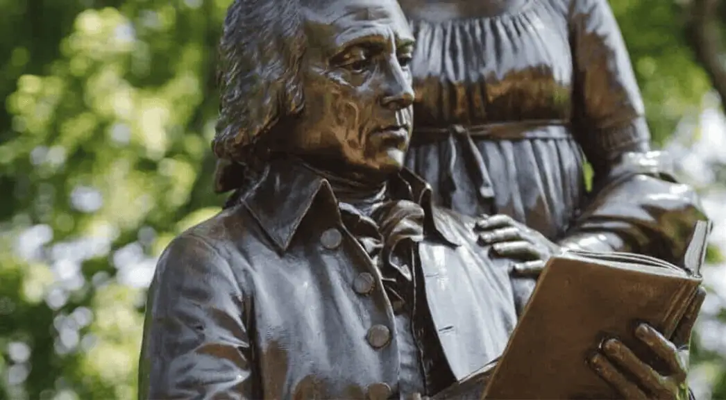 Bronze statue of James Madison in colonial-era attire reading a book, with a woman standing behind him, set in a green park setting.