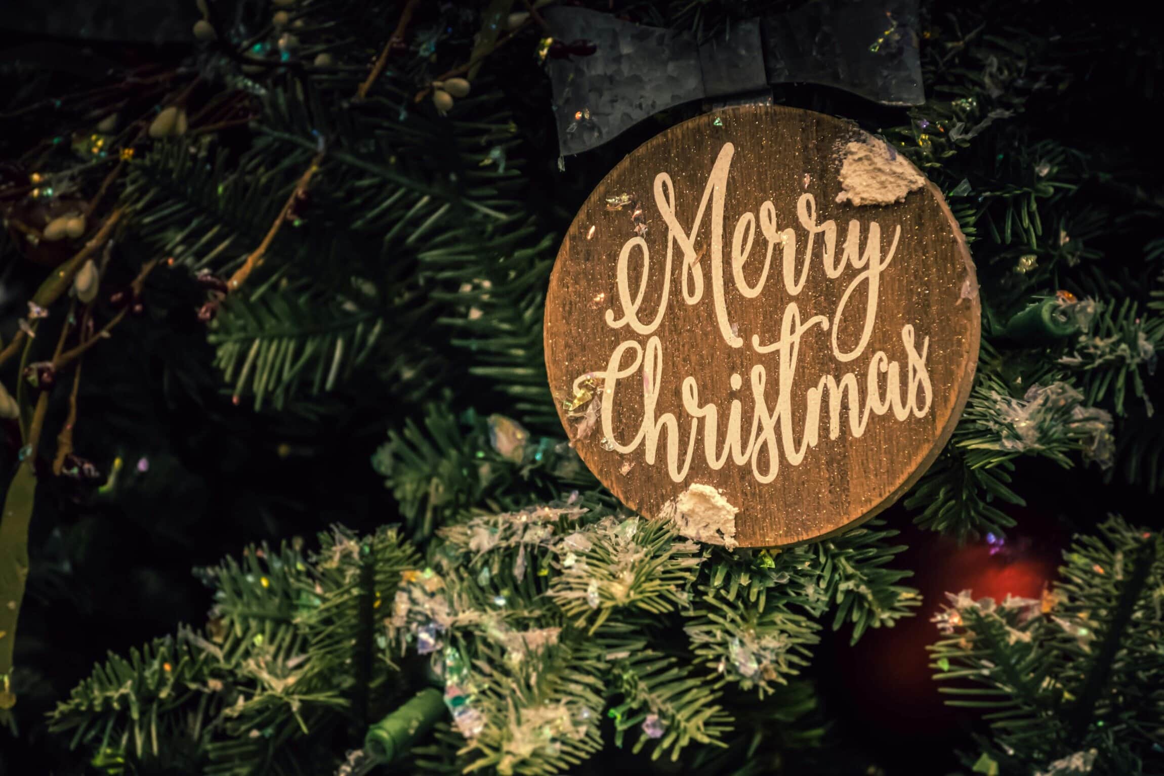 Close-up of a rustic wooden “Merry Christmas” ornament hanging on a decorated Christmas tree with evergreen branches and twinkling lights.