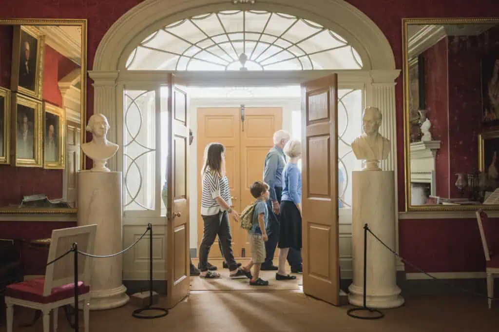 Visitors walking through the historic interior of James Madison’s Montpelier, framed by an arched doorway, portraits, and marble busts.