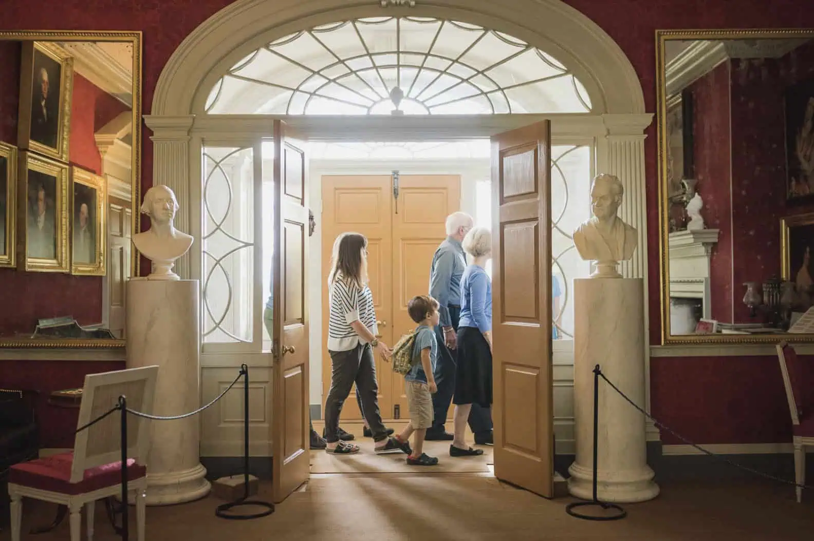 Visitors walking through the historic interior of James Madison’s Montpelier, framed by an arched doorway, portraits, and marble busts.