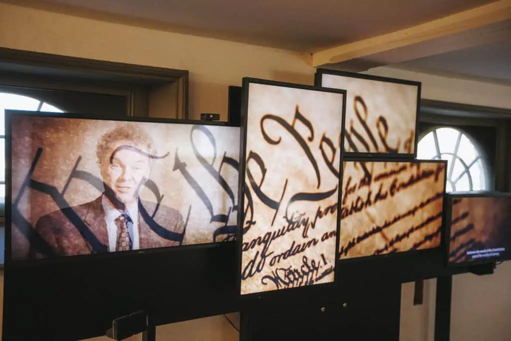 A multi-screen museum display shows a speaker alongside enlarged sections of the handwritten U.S. Constitution