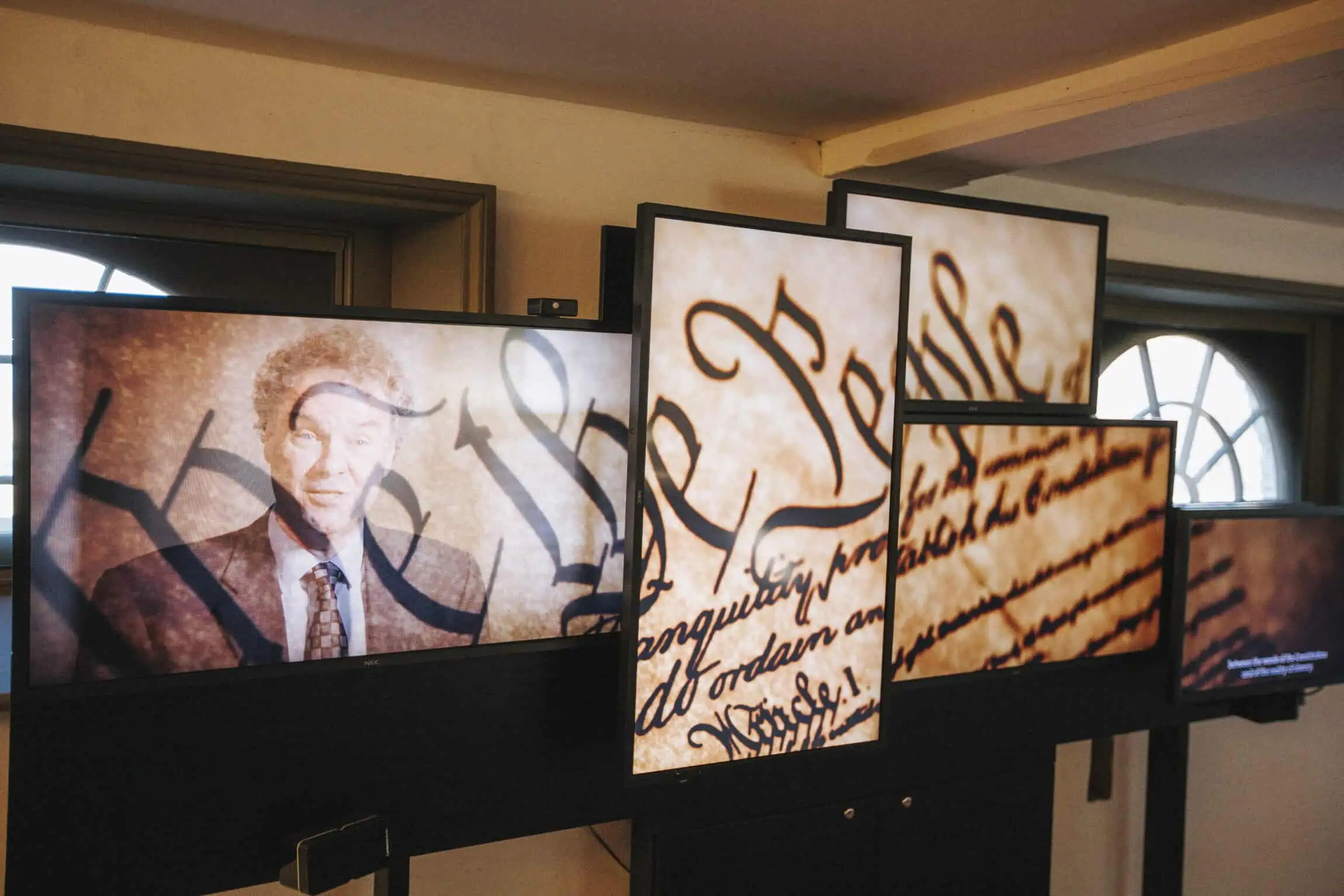 A multi-screen museum display shows a speaker alongside enlarged sections of the handwritten U.S. Constitution