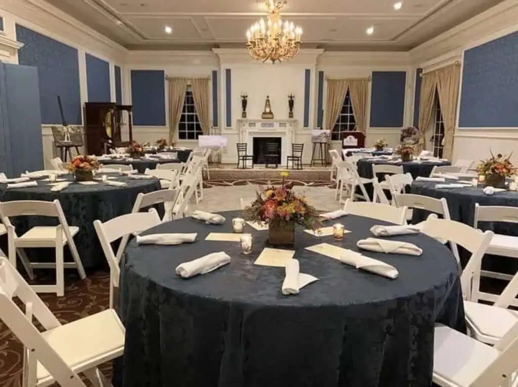 Elegant banquet dining room set with round tables, navy tablecloths, white folding chairs, floral centrepieces, candles, and formal place settings beneath a chandelier.