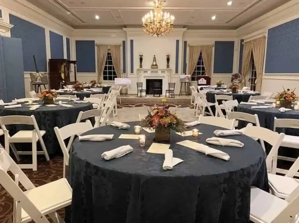 Elegant banquet dining room set with round tables, navy tablecloths, white folding chairs, floral centrepieces, candles, and formal place settings beneath a chandelier.
