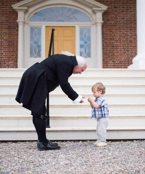 An adult dressed in historical attire bends down to greet a toddler in casual clothes in front of a large building with columns and steps.
