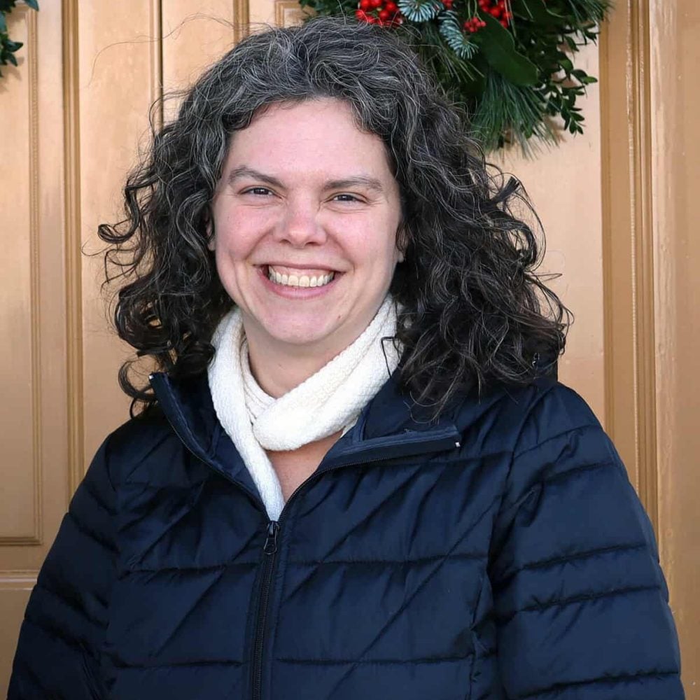 A woman with curly gray hair wearing a navy blue jacket and white scarf smiles in front of a beige door decorated with a wreath.